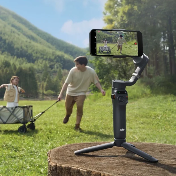 Черный стабилизатор DJI Osmo Mobile 6 на комплектном настольном триподе на открытой зеленой поляне на фоне гор удерживает смартфон со сценой бегущих детей